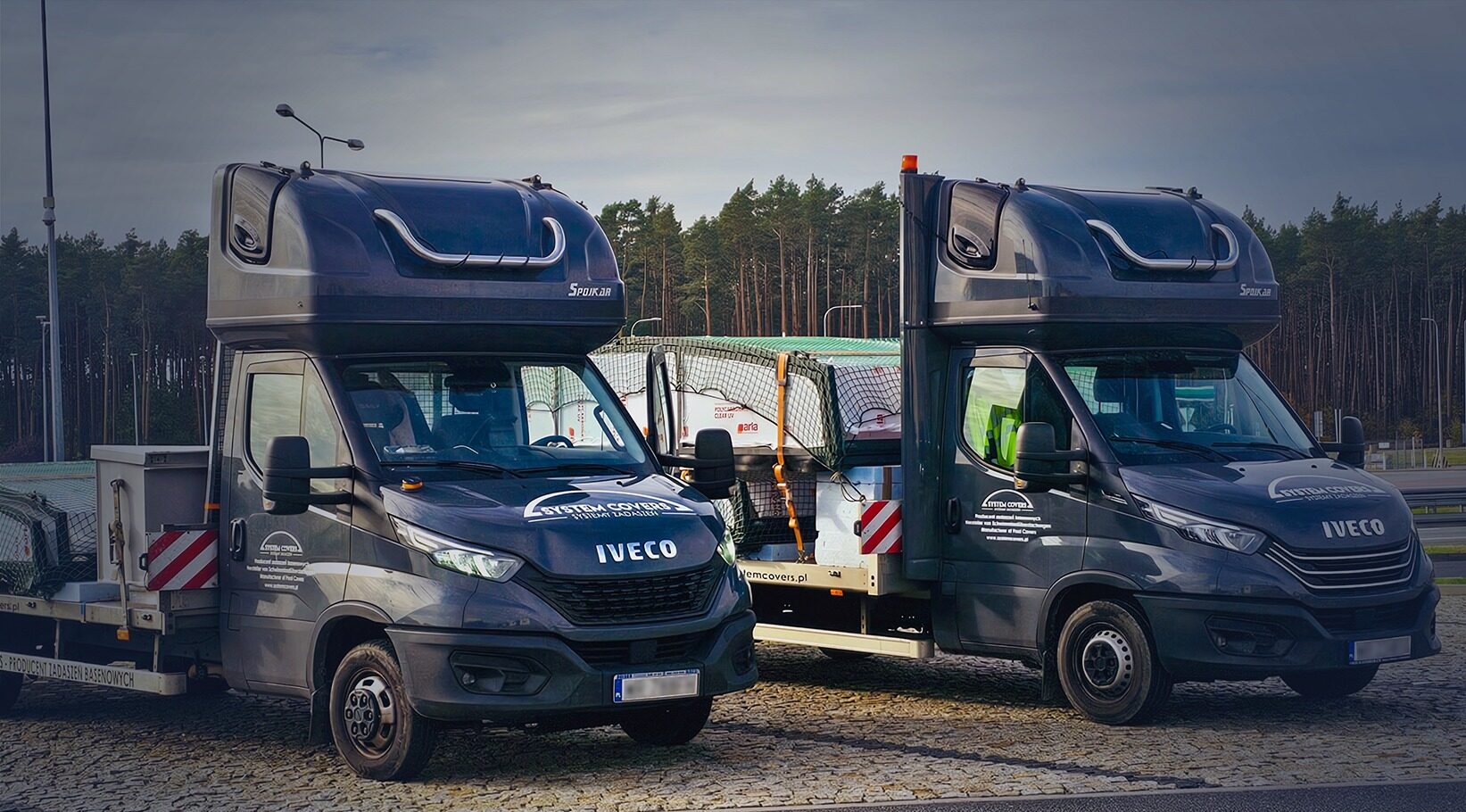 Dwa pojazdy dostawcze Iveco z przyczepami załadowanymi elementami zadaszeń, w tle las.