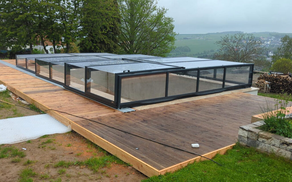 Zadaszenie basenu na drewnianym tarasie w otoczeniu zieleni i wzgórz w tle.