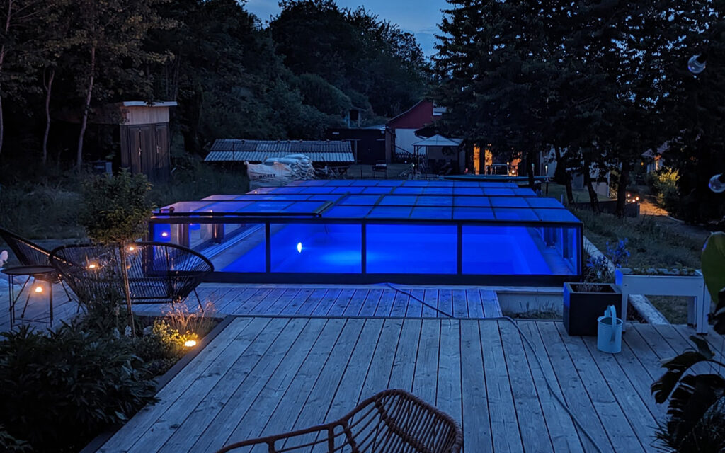 Podświetlony basen z nowoczesnym zadaszeniem w ogrodzie nocą.
