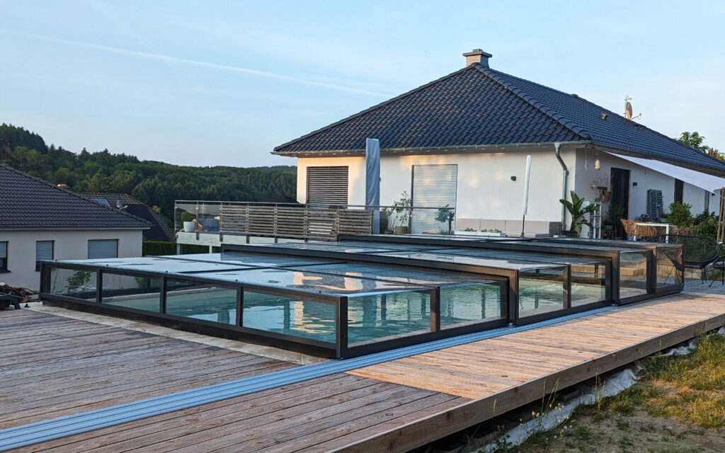 Basen z przesuwnym zadaszeniem na drewnianym tarasie, w tle dom jednorodzinny z ogrodem.