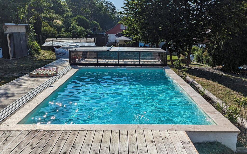 Basen z zamontowanym przesuwnym zadaszeniem, otoczony zielenią i drewnianym tarasem.