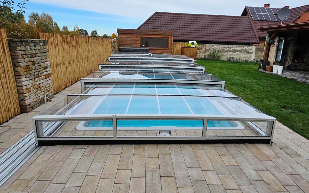 Zadaszenie basenu z przezroczystymi panelami w ogrodzie, otoczone drewnianym płotem i zieloną trawą.