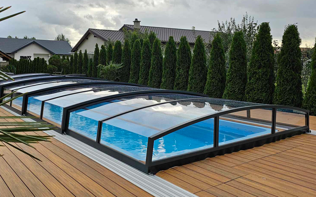 Niskie zadaszenie basenowe na drewnianym tarasie, otoczone zielonym żywopłotem i domami w tle.