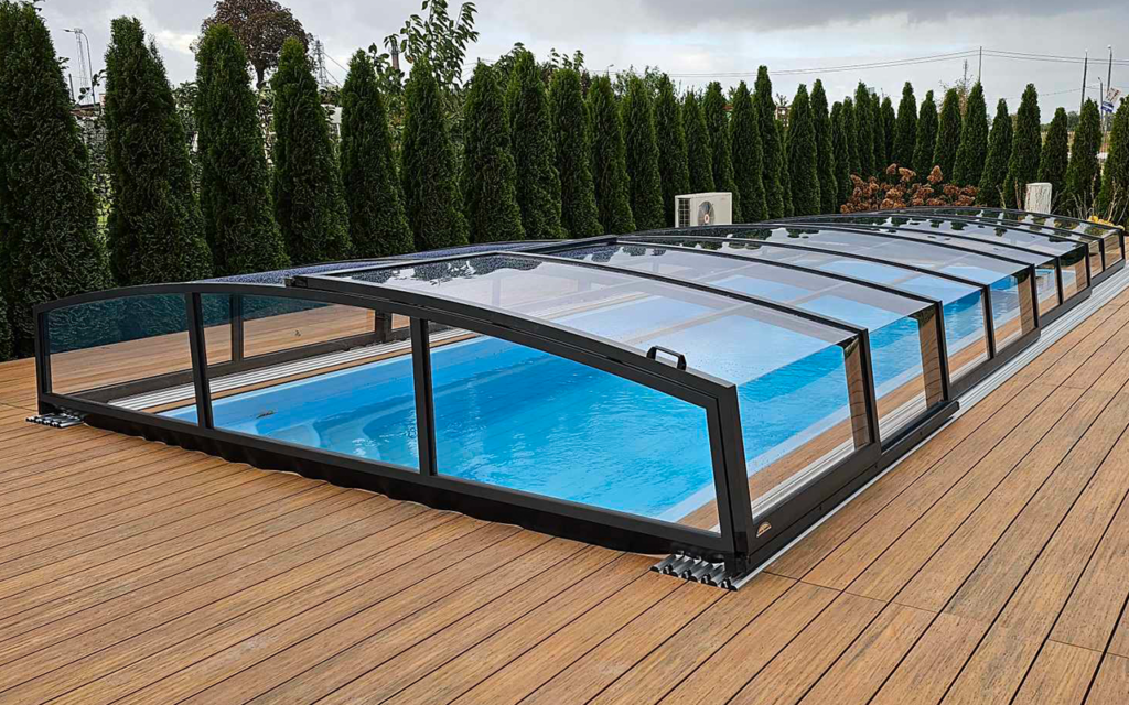 Nowoczesna zadaszona konstrukcja basenowa z przeźroczystymi panelami i drewnianym tarasem, otoczona wysokimi tujami.