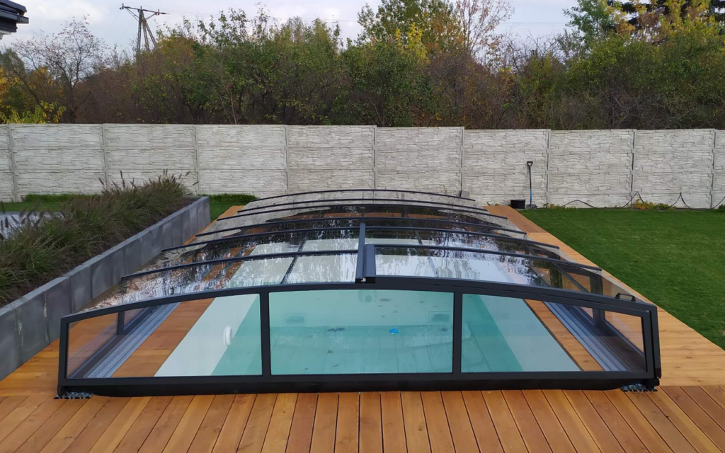 Zadaszenie basenu na drewnianym tarasie w ogrodzie, otoczone zielenią i białym ogrodzeniem.