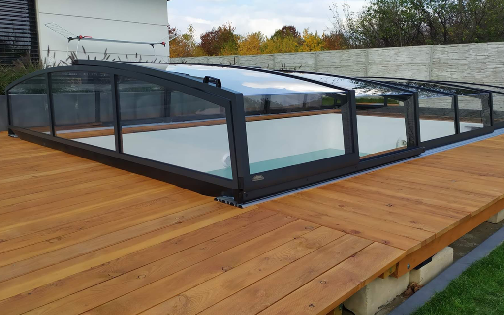 Pokrycie basenu na drewnianym tarasie, przezroczyste panele z ciemnymi ramami, w tle roślinność i mur.