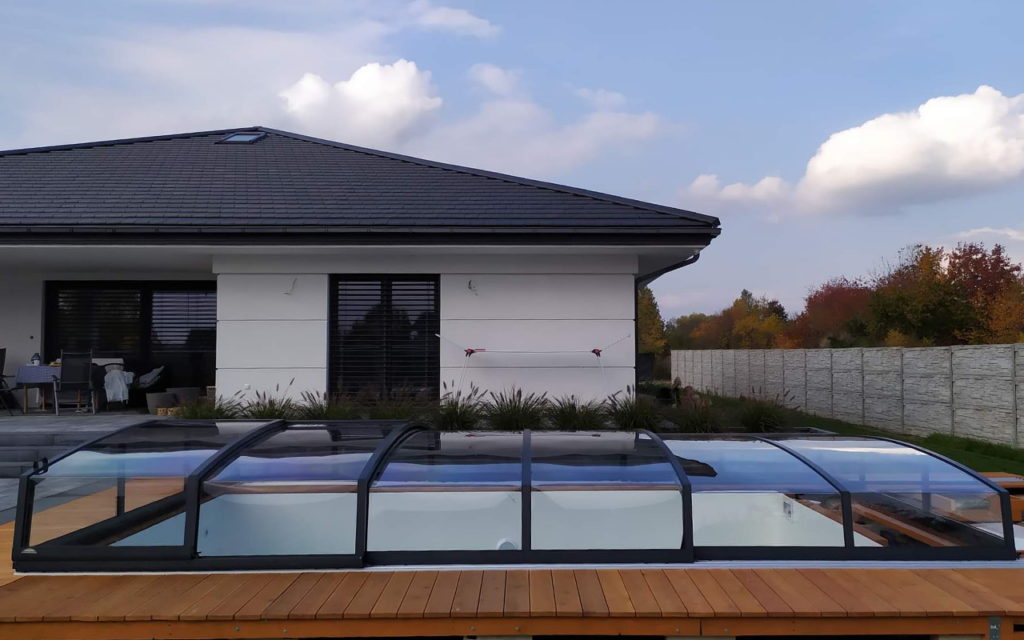 Zabudowa basenowa przy nowoczesnym domu, ogrodzona drewnianym tarasem, w tle niebo z chmurami i roślinność.