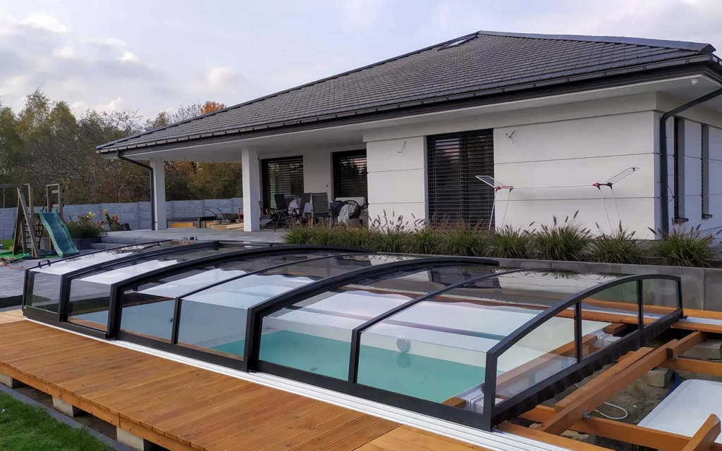 Kryte zadaszenie basenu obok nowoczesnego domu z tarasem, otoczone roślinnością i ogrodem.