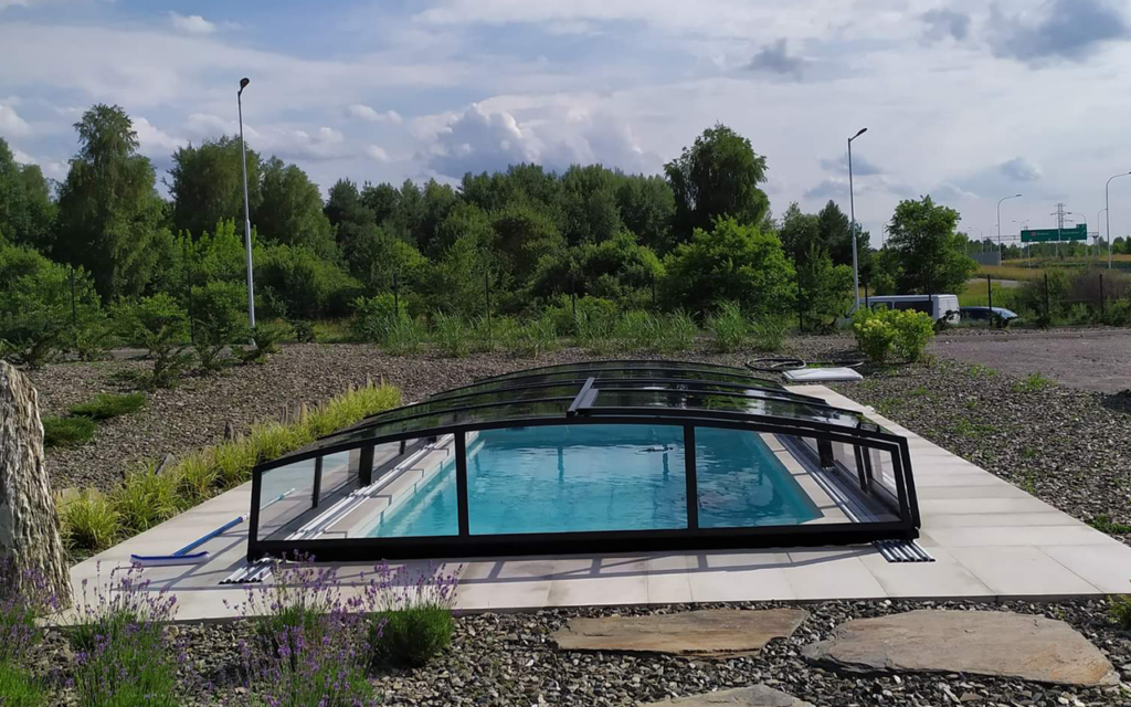 Basen z zadaszeniem, otoczony zielenią, na tle drzew i jasnego nieba.