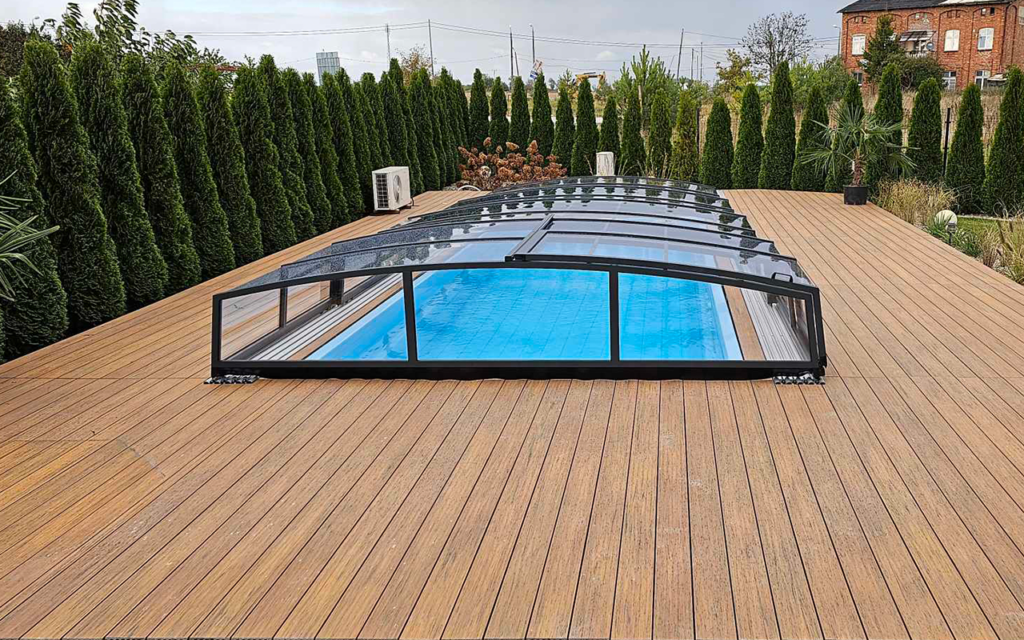 Basen kryty osłoną na drewnianym tarasie otoczony wysokimi krzewami.