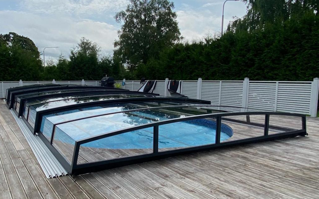Basen z nowoczesnym zadaszeniem aluminiowym na drewnianym tarasie, otoczony zielenią i ogrodzeniem.
