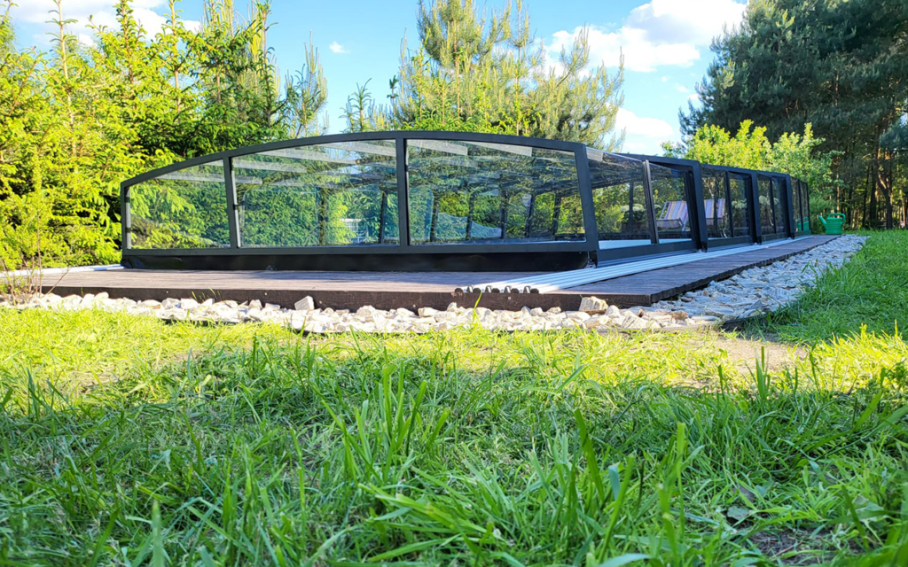 Przeszklona osłona basenowa w ogrodzie otoczona zielenią i drzewami iglastymi.