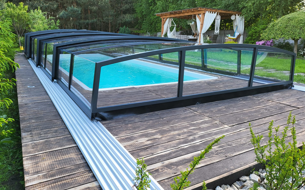 Zadaszenie basenowe z czarną aluminiową ramą na drewnianym tarasie, w tle altana i zieleń.