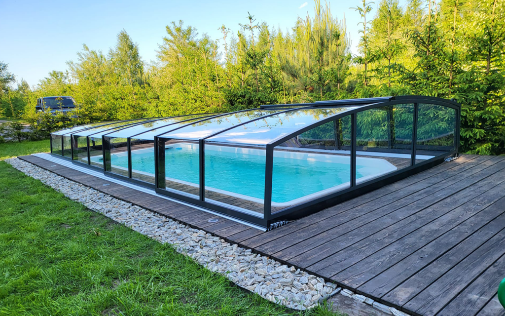 Basen z nowoczesnym przeszklonym zadaszeniem na drewnianym tarasie, otoczony zielenią.