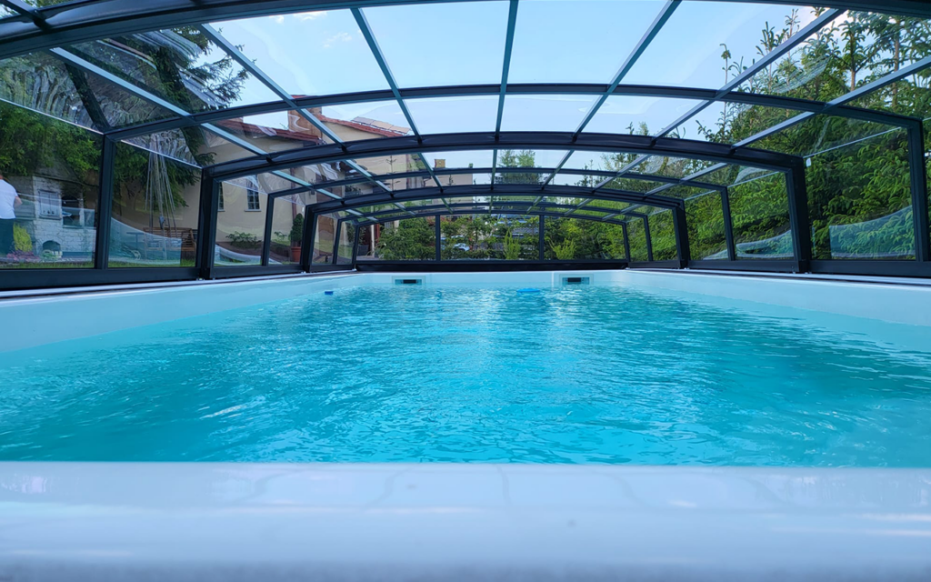 Zadaszony basen z przezroczystym dachem w otoczeniu zieleni i budynków.