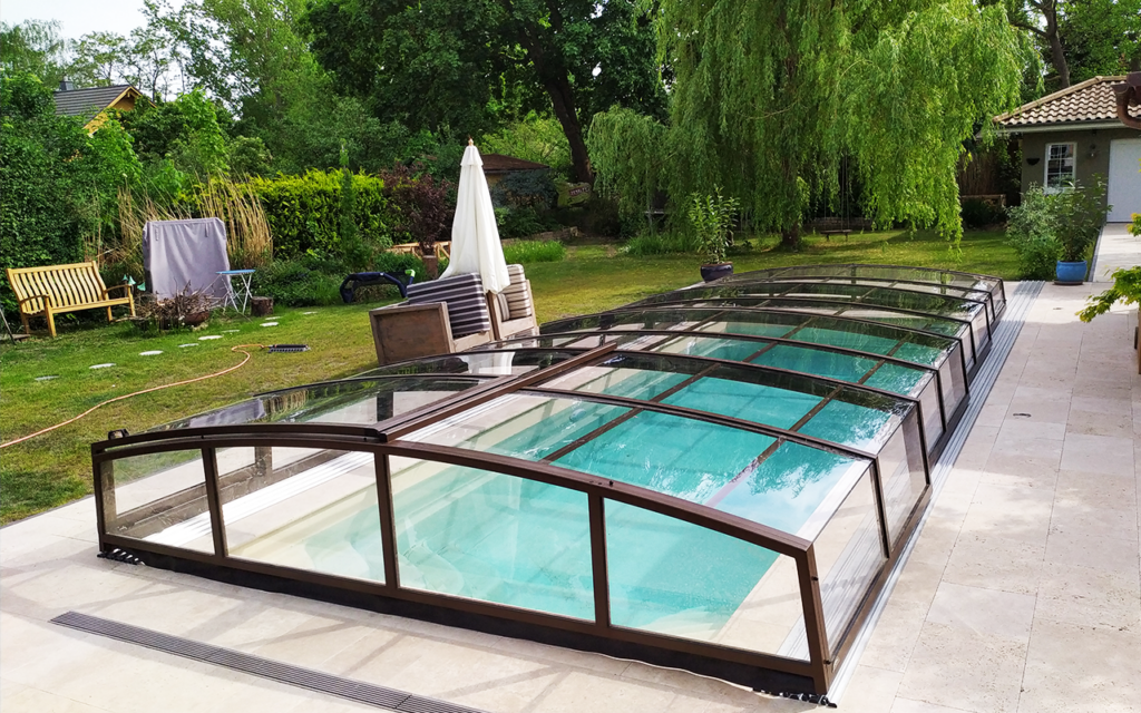Przeszklona osłona basenu w ogrodzie, otoczona zielenią i meblami ogrodowymi.