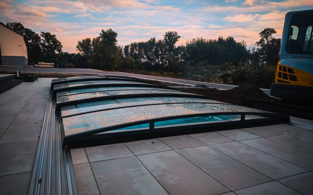 Zadaszenie basenu z przezroczystymi panelami na dużym tarasie, obok widoczna koparka na tle zieleni.