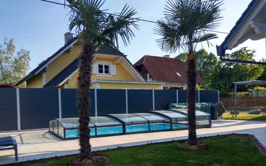 Basen ogrodowy z osłoną w słonecznym ogrodzie, otoczony palmami i nowoczesnym ogrodzeniem.