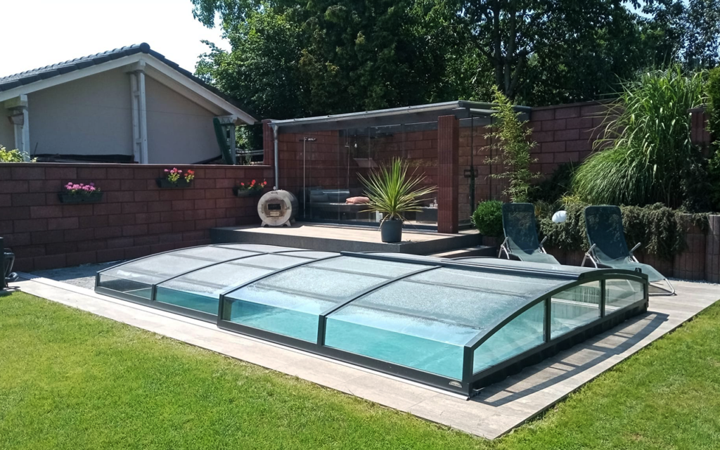 Przeszklona osłona basenowa na zielonym trawniku, w tle meble ogrodowe i rośliny.