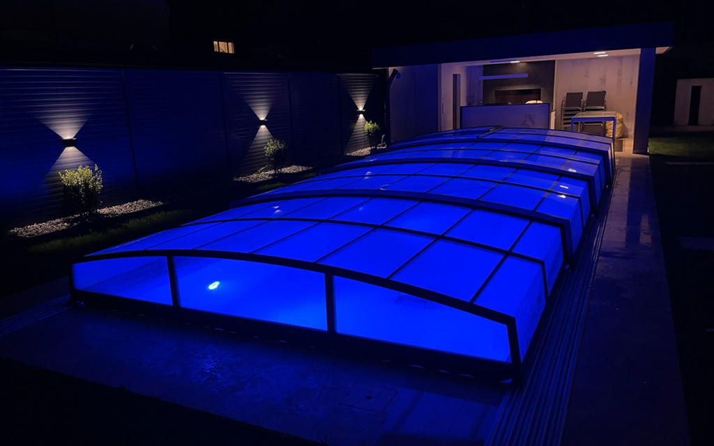 Oświetlone, nowoczesne zadaszenie basenu nocą z niebieskim podświetleniem, w tle altana ogrodowa.