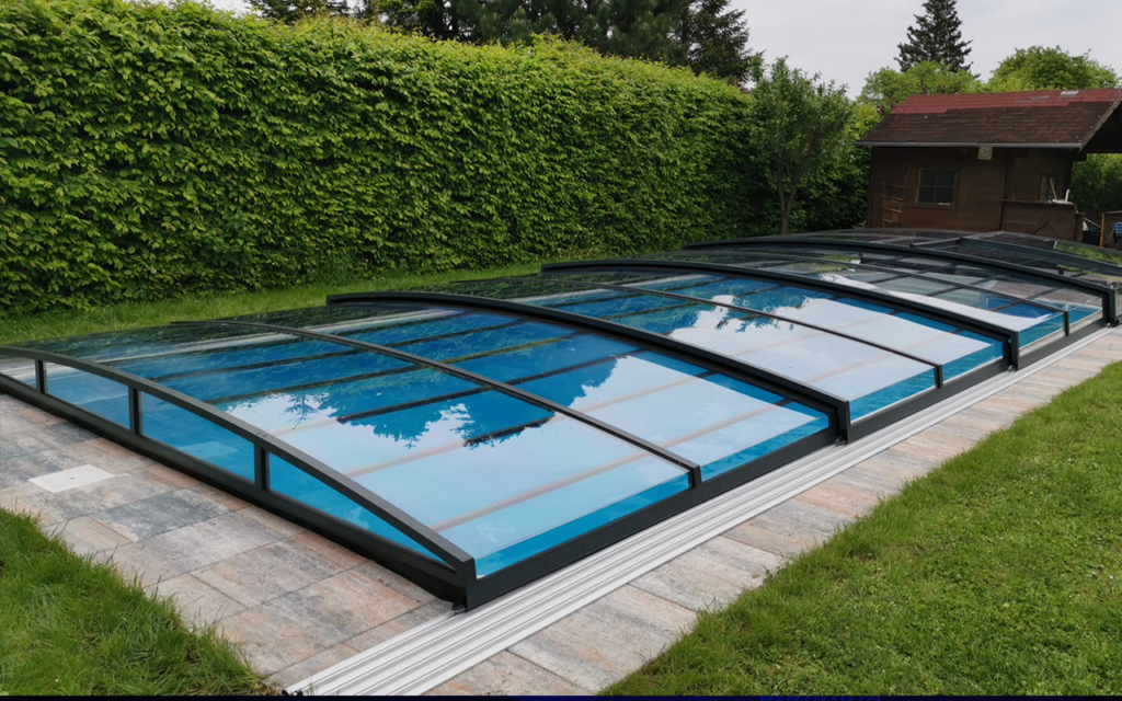 Basen z zadaszeniem w ogrodzie otoczony zielenią, zbudowanym z przezroczystych paneli i metalowych ram.