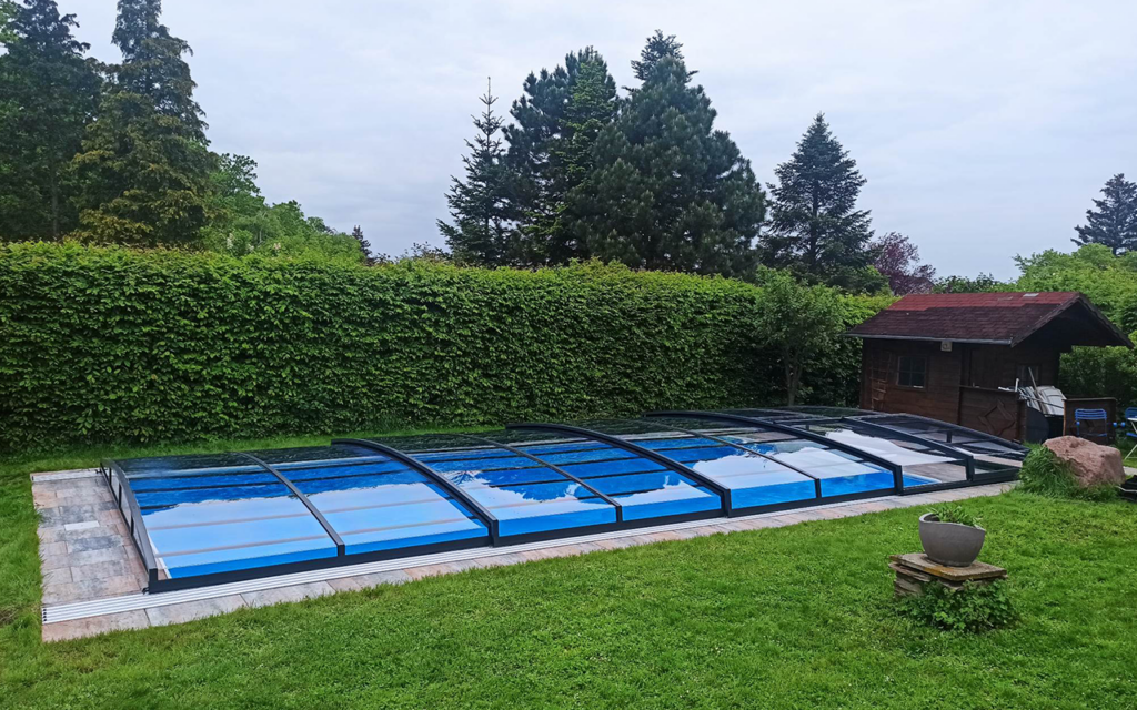 Zadaszenie basenu w ogrodzie otoczone zielenią i drzewami, obok drewniany domek i donica na trawie.