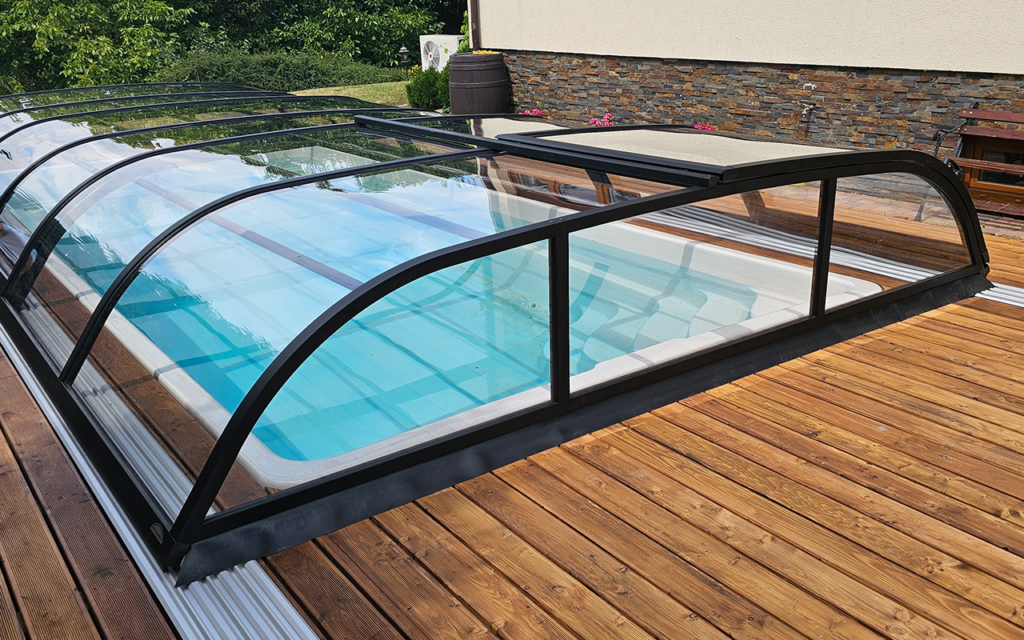 Zadaszenie basenowe z przezroczystymi panelami na drewnianym tarasie, w tle zielone drzewa.
