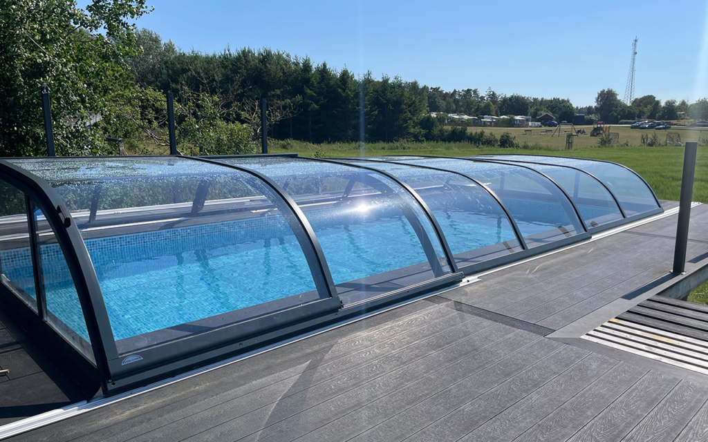Przeszklona zabudowa basenu na drewnianym tarasie, w tle drzewa i trawnik.