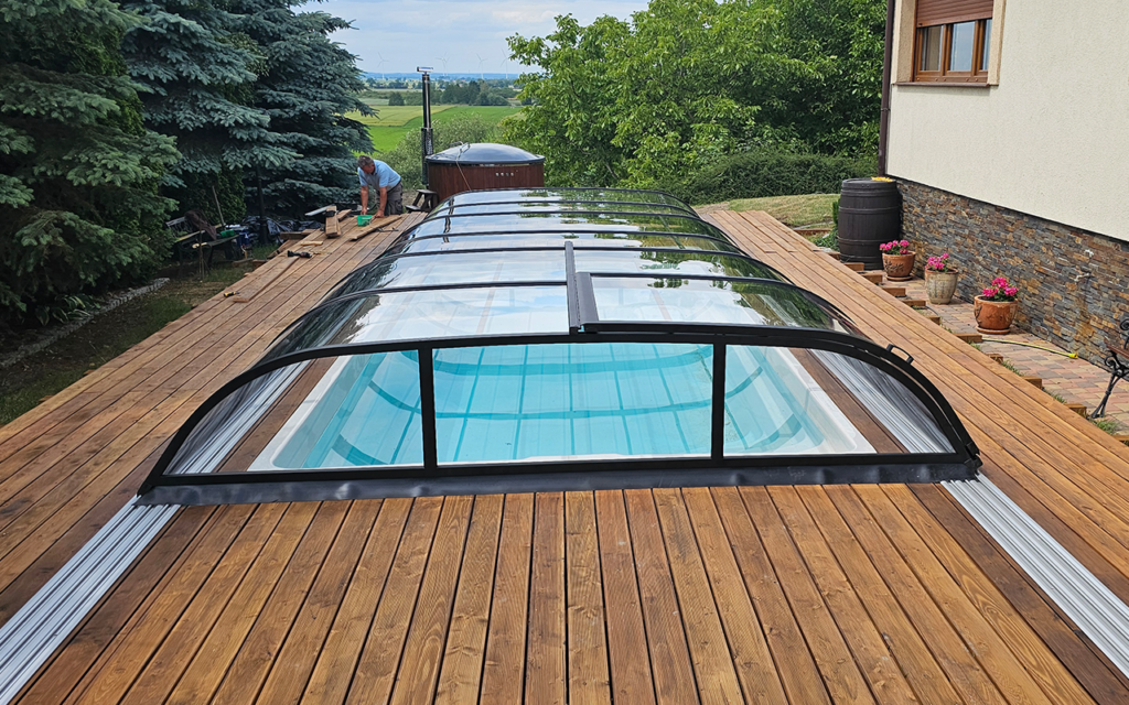 Basen z osłoną wykonaną z przezroczystych paneli, otoczony drewnianym tarasem, w tle zieleń i drzewa.