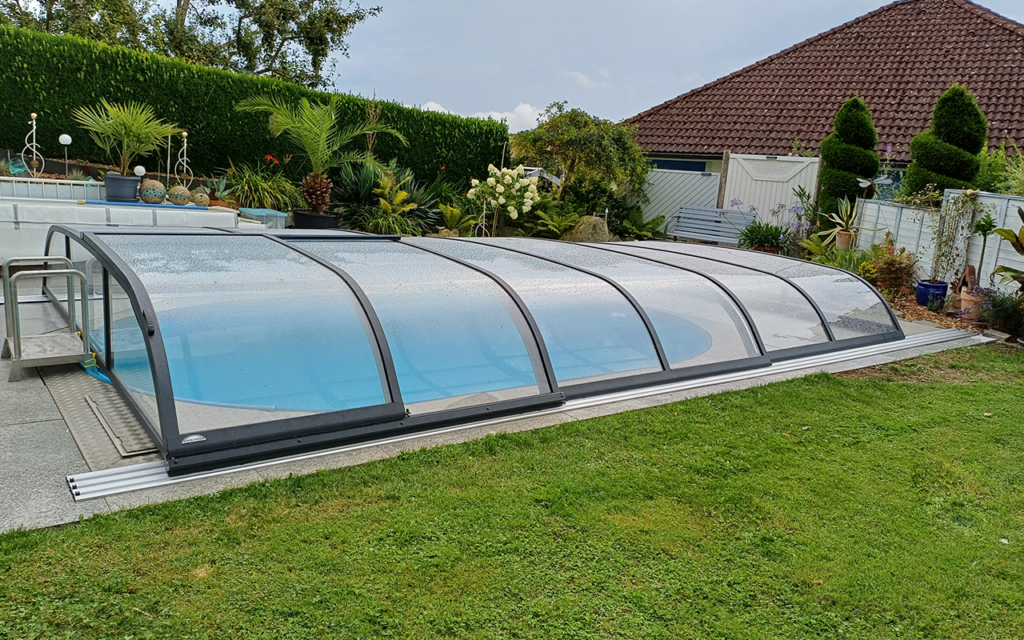 Basen z przezroczystym zadaszeniem w ogrodzie, otoczony roślinnością i trawnikiem.