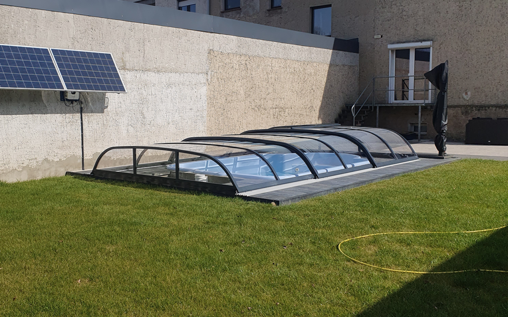 Zadaszenie basenu na trawniku, obok znajduje się panel słoneczny na ścianie budynku.