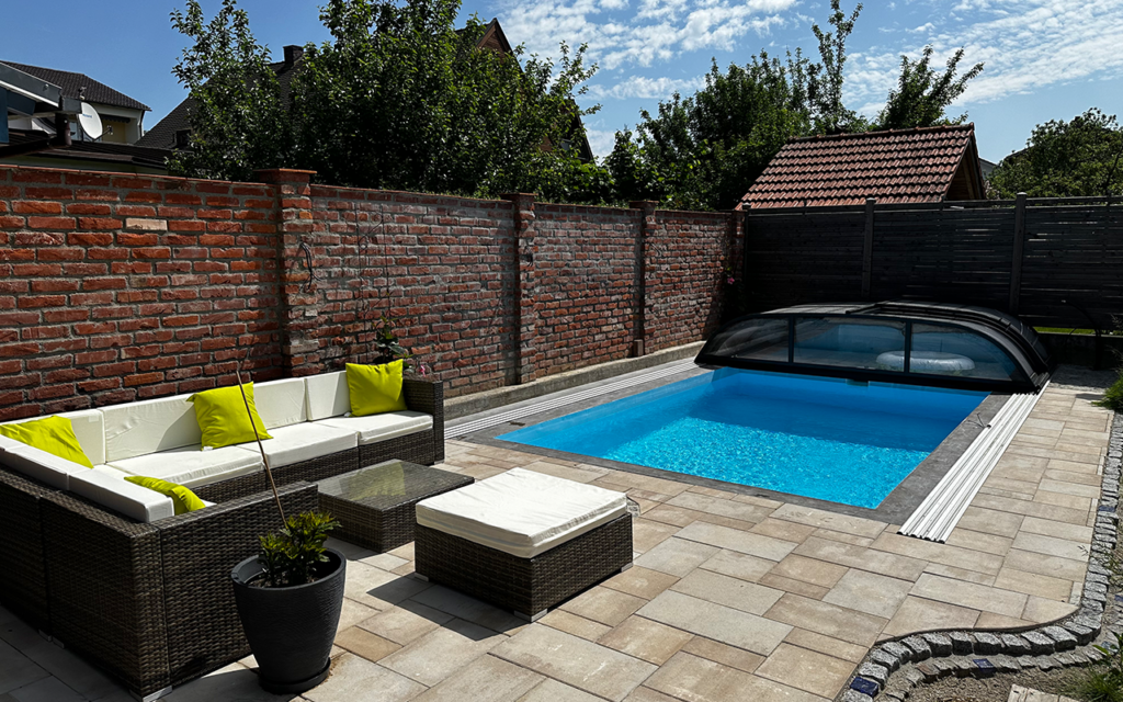 Basen z zadaszeniem w ogrodzie, otoczony rattanowymi meblami, przy ceglanej ścianie i zieleni.