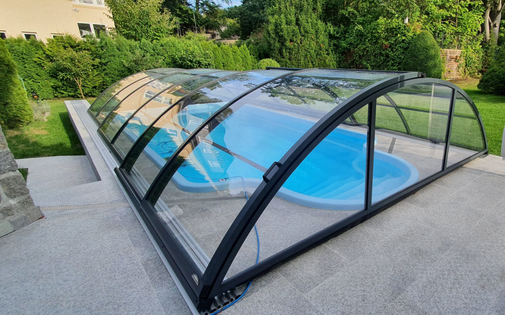 Zadaszenie basenu z przezroczystymi panelami na tle ogrodu z drzewami i krzewami.