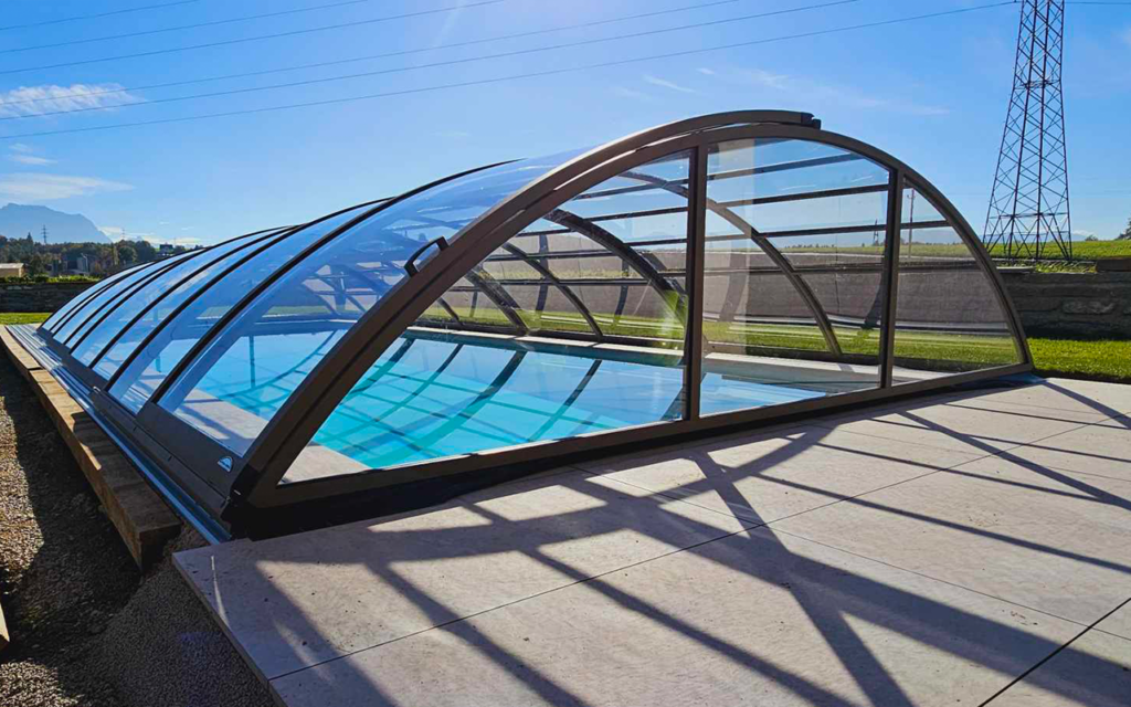 Basen z przezroczystym, łukowatym zadaszeniem na tle zielonego krajobrazu i słonecznego nieba.