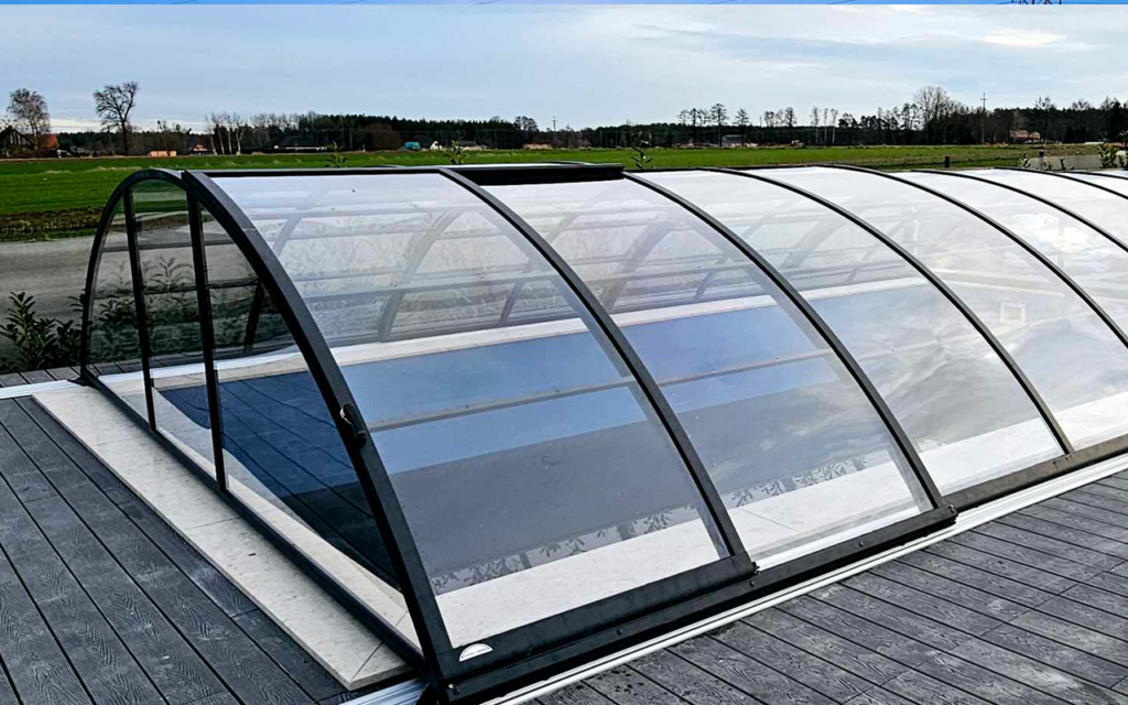 Przeszklona zabudowa basenu na drewnianym tarasie, w tle zielone pola i drzewa.