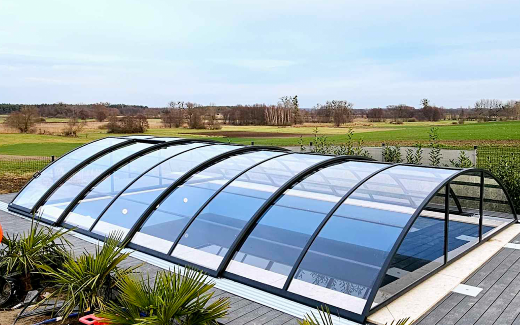 Przeszklona zabudowa basenowa otoczona zielenią, w tle otwarta przestrzeń z polami i drzewami.