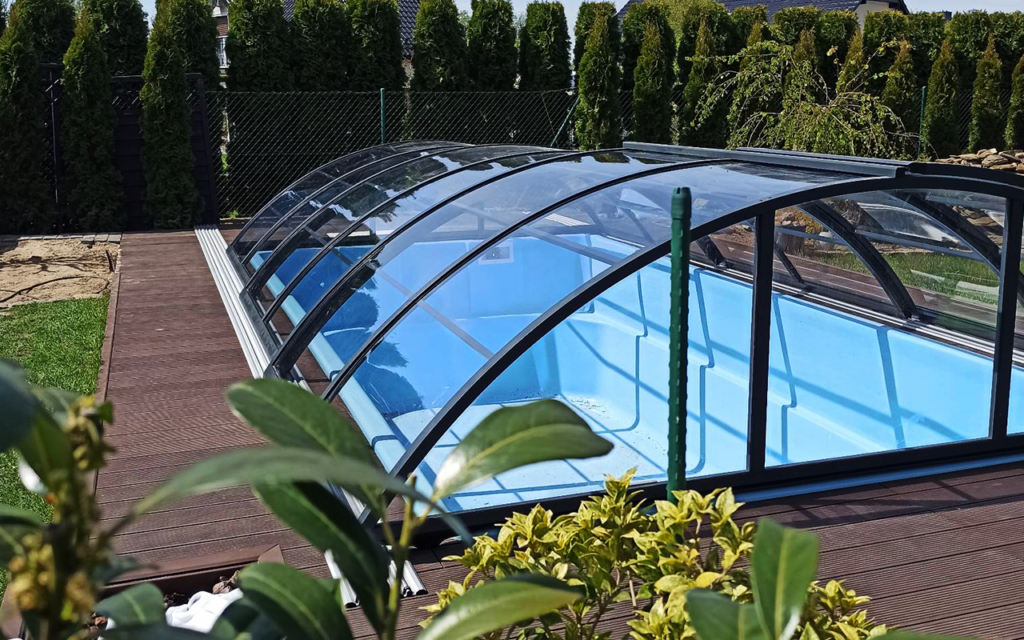 Zadaszenie basenu w ogrodzie, przezroczysta konstrukcja z metalu i szkła otoczona zielenią.