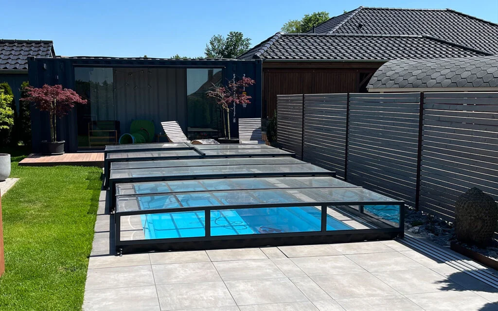 Basen z przezroczystym zadaszeniem na przydomowym podwórku, otoczony nowoczesnym ogrodzeniem i zielenią.