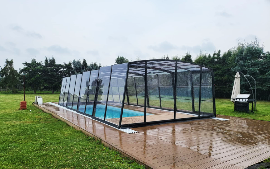 Nowoczesne zadaszenie basenowe na drewnianym tarasie w otoczeniu zieleni.