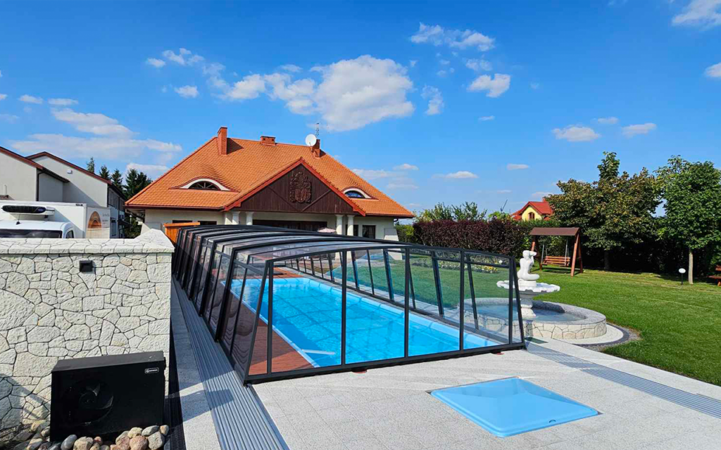 Zadaszenie basenu przy domu jednorodzinnym, jasne niebo, ogród z altaną i fontanną w tle.