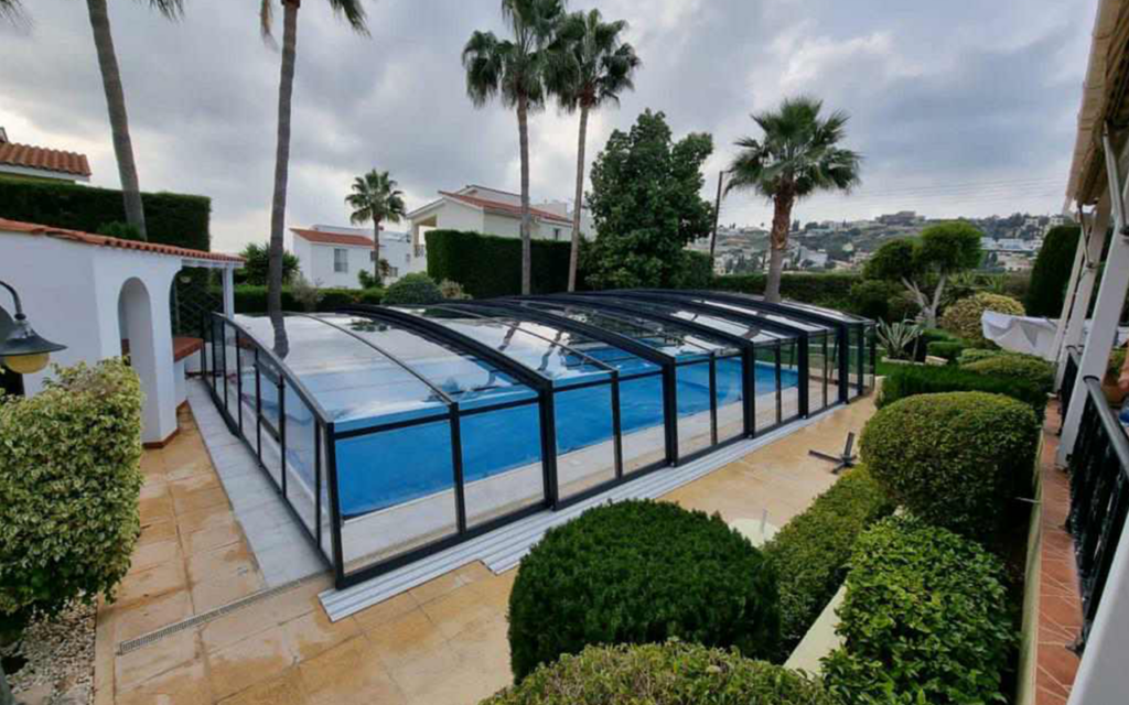 Zadaszenie basenu w ogrodzie z palmami i krzewami, w tle budynki i pagórkowaty krajobraz.