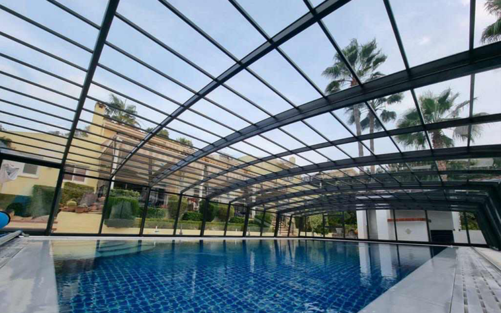 Basen z niebieską mozaiką i zadaszeniem z przesuwanej konstrukcji, otoczony palmami.